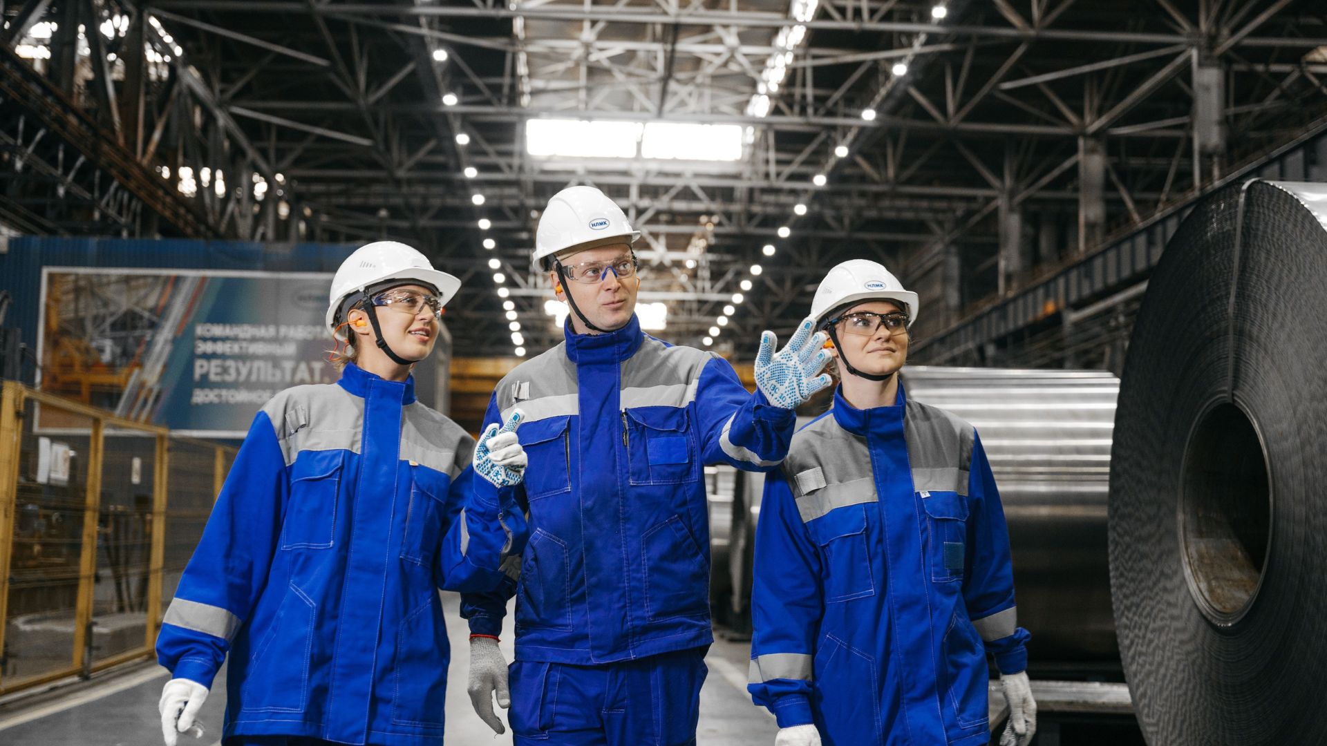 Duas mulheres e um homem com uniforme azul e capacetes brancos em uma fábrica