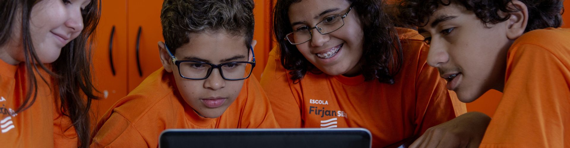 quatro estudantes com uniforme laranja da escola Firjan SESI olhando para um notebook