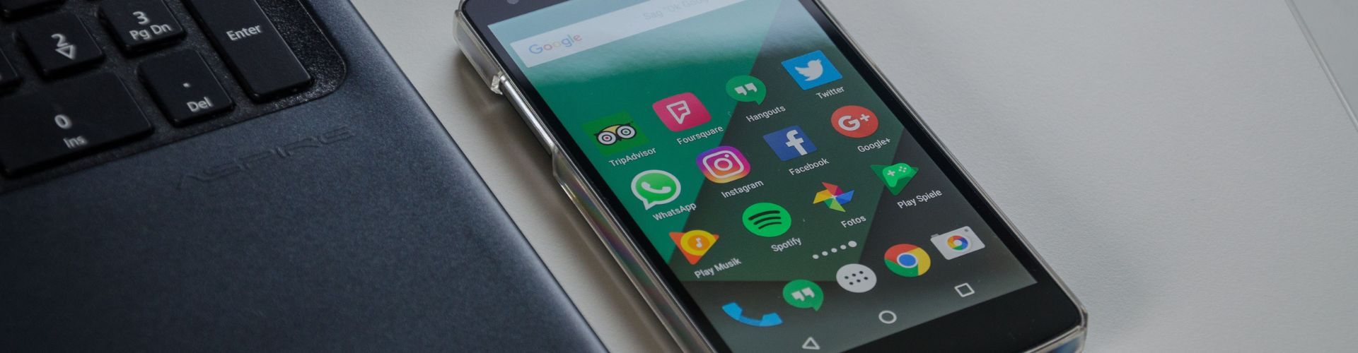 imagem exibe parte pequena de um notebook, com algumas teclas, e um celular ao lado, com ícones de redes sociais e outros aplicativos