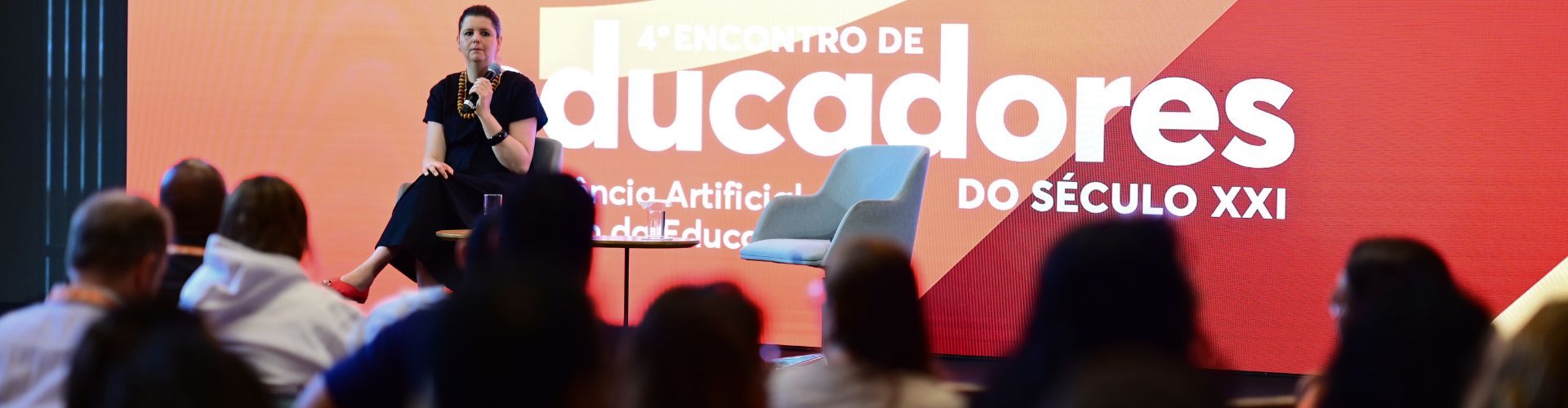 Fotografia em plano médio de um evento presencial. Em primeiro plano, desfocadas, veem-se as cabeças e costas das pessoas que compõem a plateia. Ao fundo, em destaque iluminado por um grande telão, está o palco.