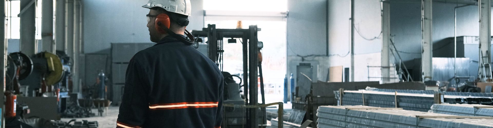 Um trabalhador de costas usa capacete branco, protetor auricular laranja e uniforme com faixas refletivas enquanto observa o interior de um amplo galpão industrial equipado com empilhadeira e materiais de construção.