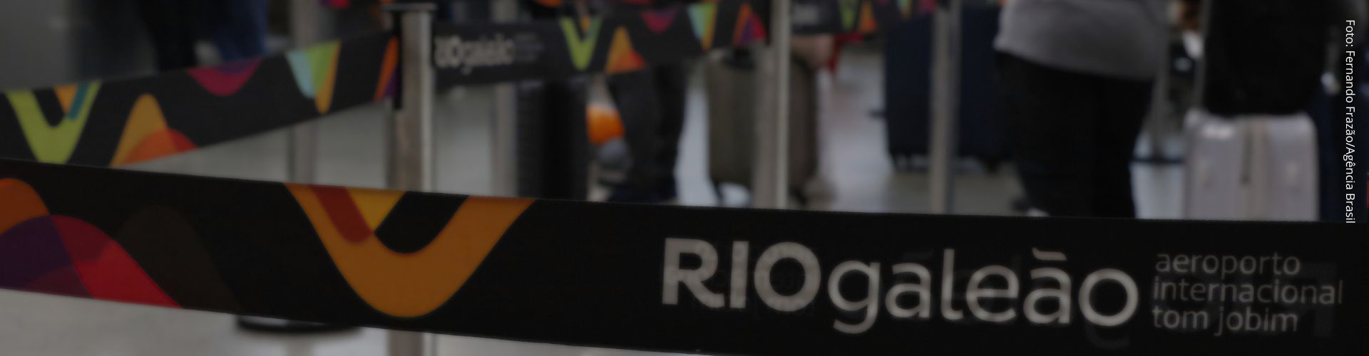 faixa com o texto RIOGaleão com filas de passageiros desfocada ao fundo