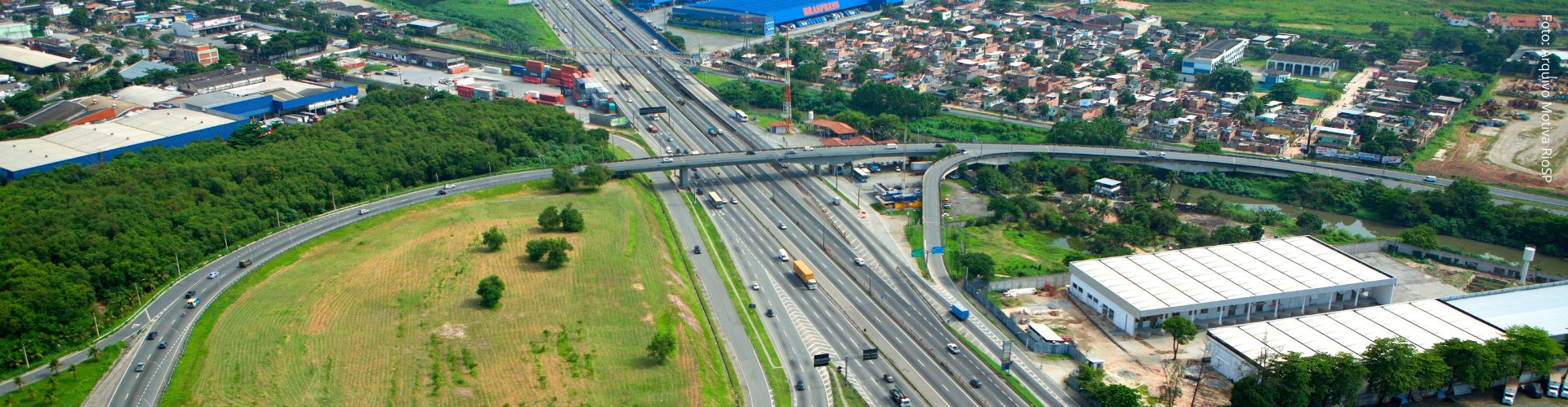 Vista aérea de complexo viário com rodovia movimentada, viadutos, galpões industriais e área urbana ao fundo.   