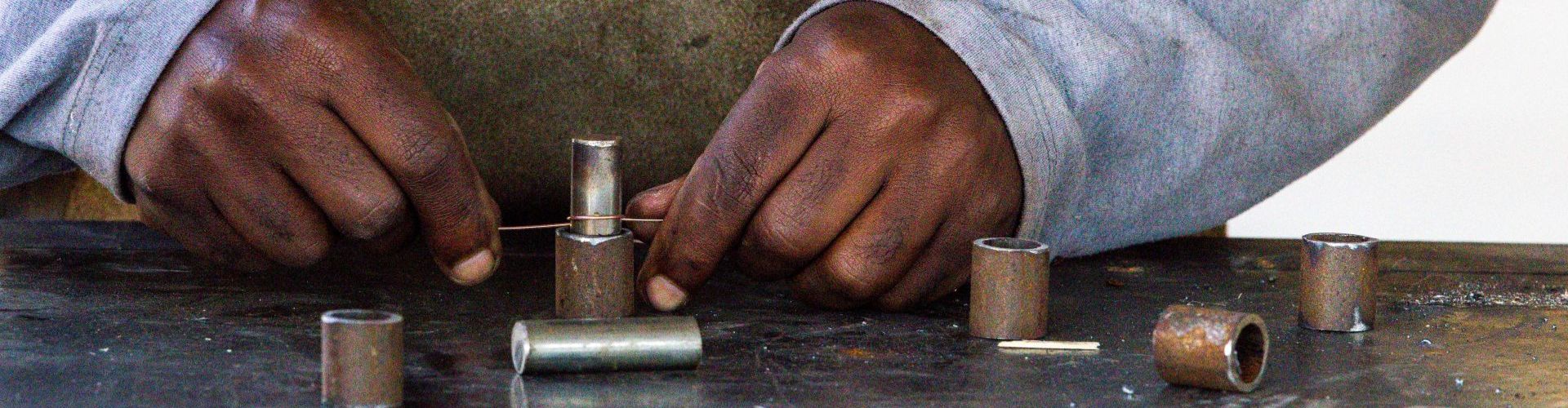 Mãos de um artesão ajustando um fio metálico ao redor de pequenos cilindros de metal sobre uma bancada