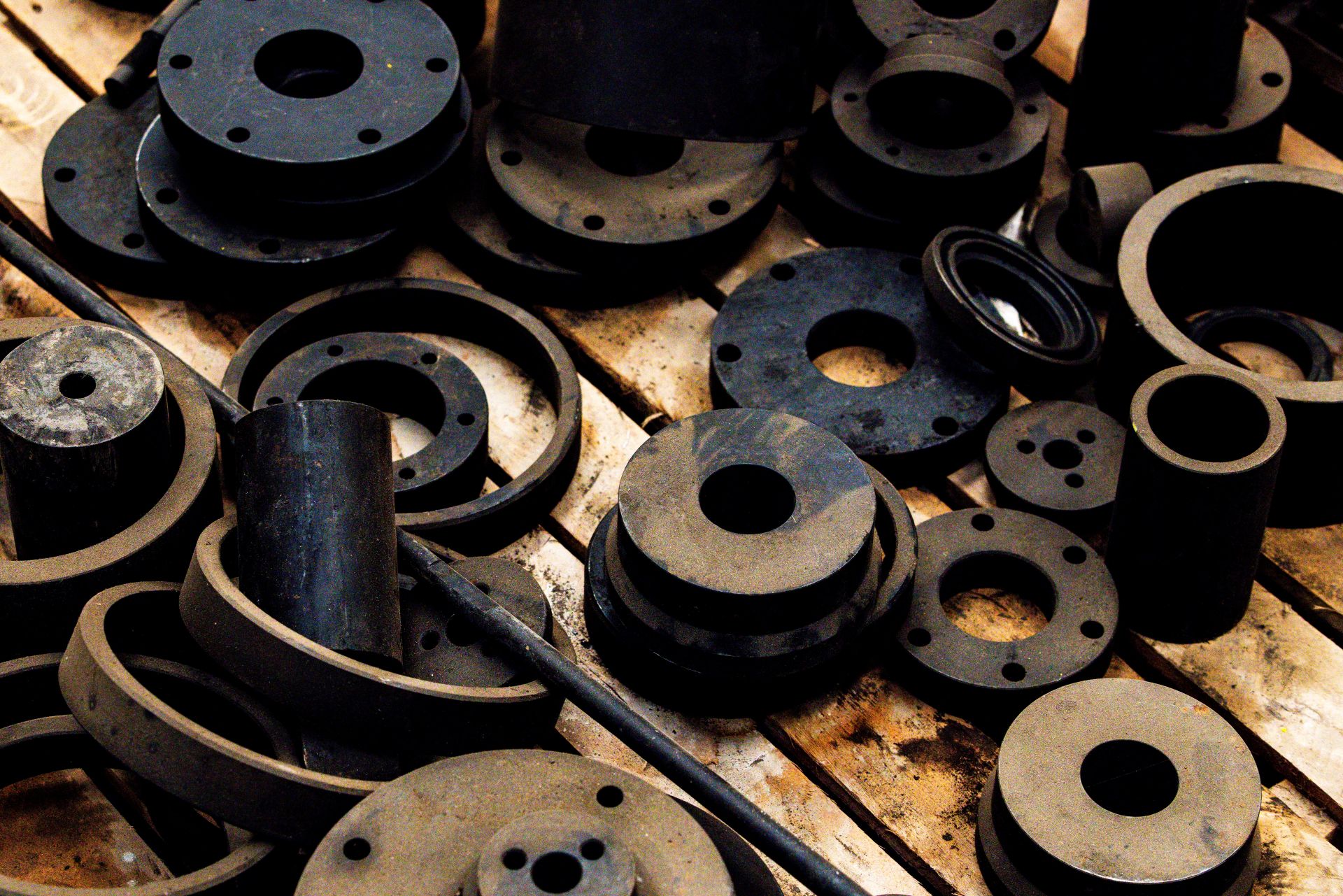 Várias peças industriais e flanges metálicas pretas e cilíndricas espalhadas sobre um pallet de madeira.
