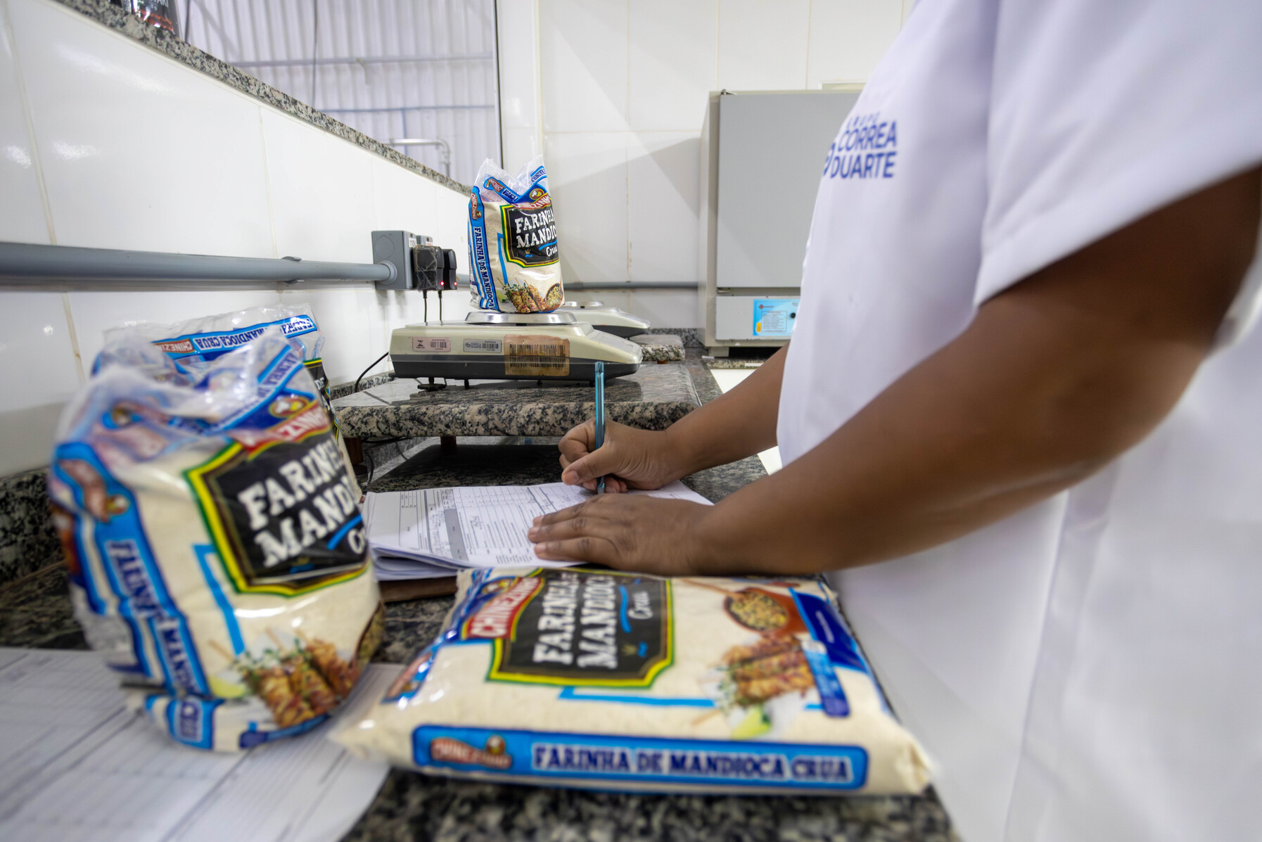Trabalhadora regista dados num documento junto a pacotes de farinha de mandioca e balança de precisão.
