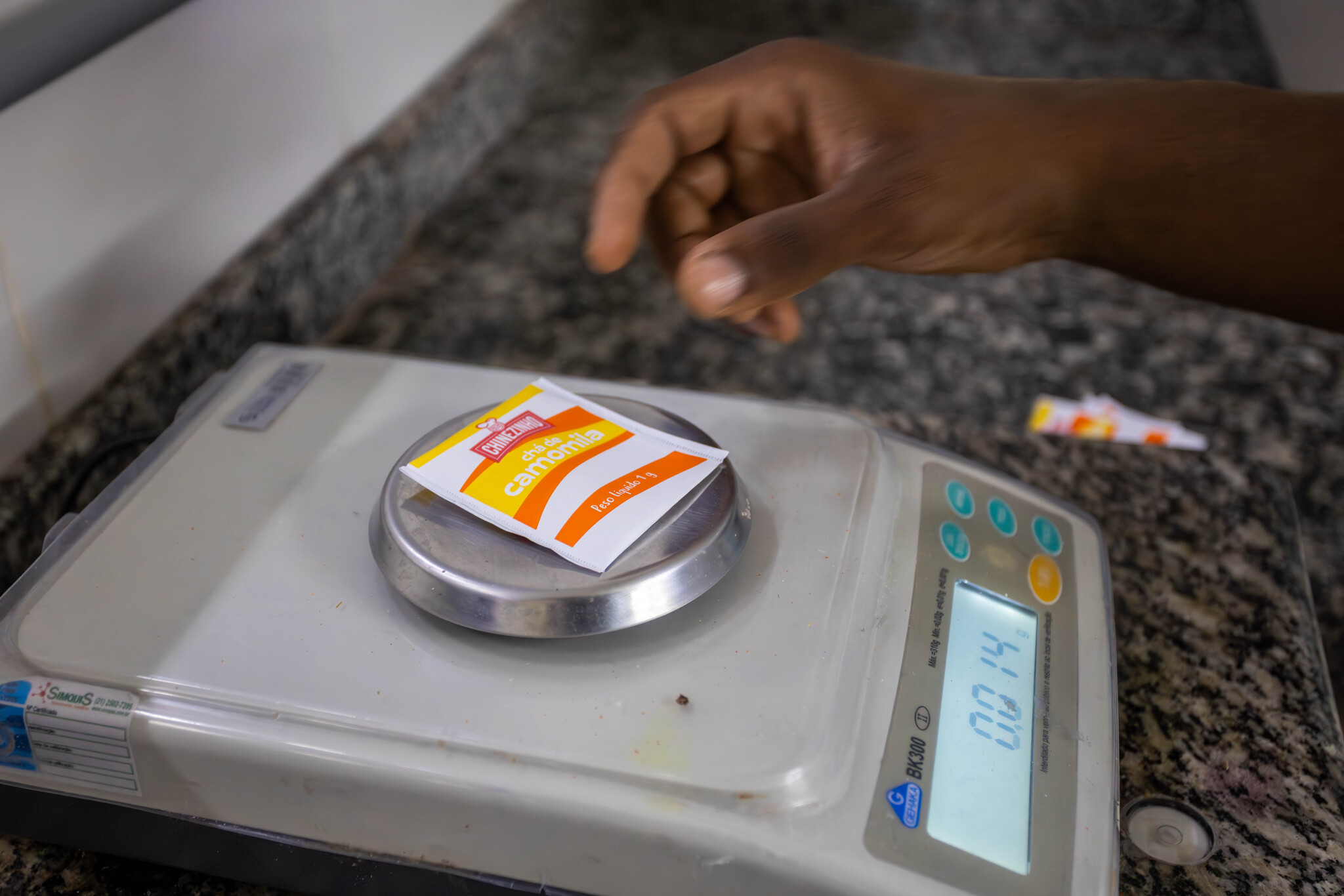 Mão de pessoa a pesar uma saqueta de chá de camomila numa balança digital de precisão.