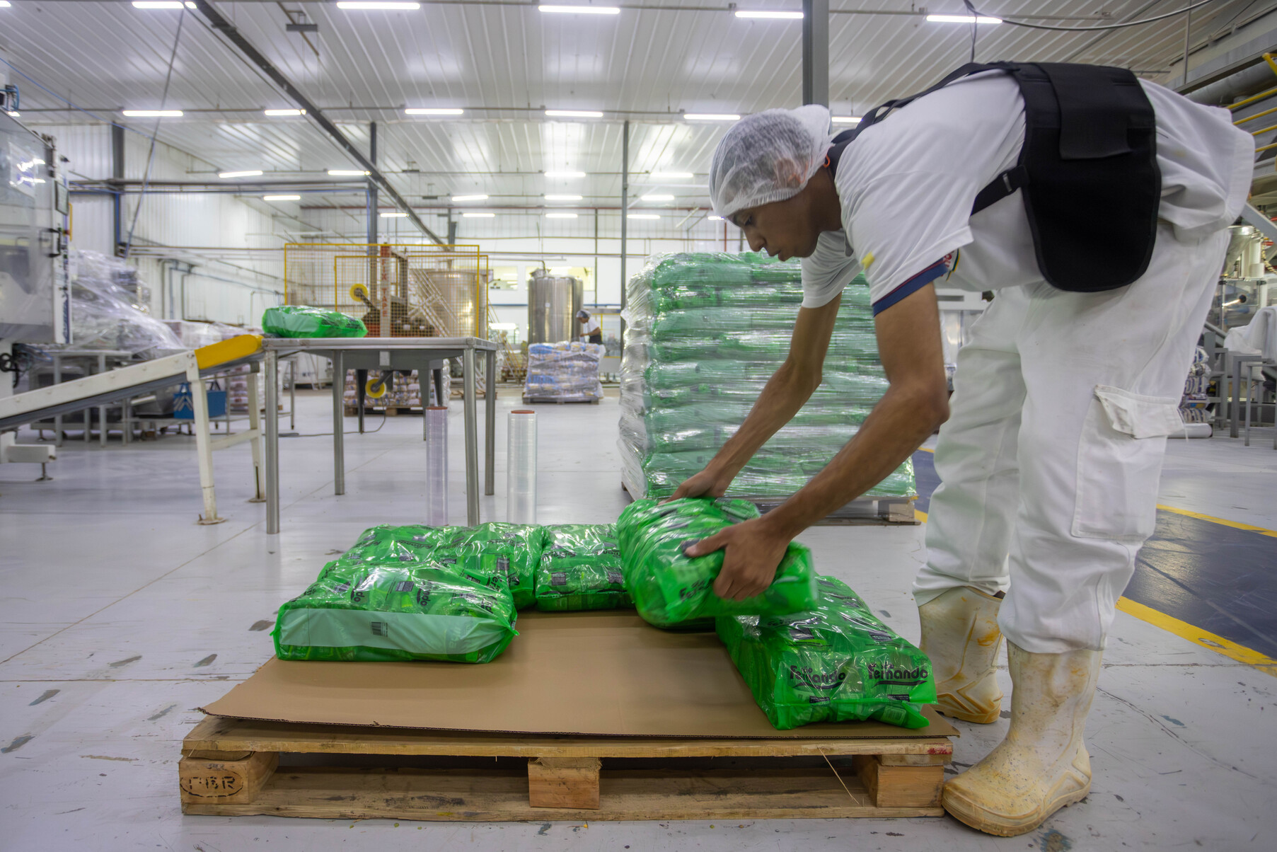 Trabalhador uniforme de branco empilha pacotes verdes de alimentos sobre um palete de madeira na fábrica.