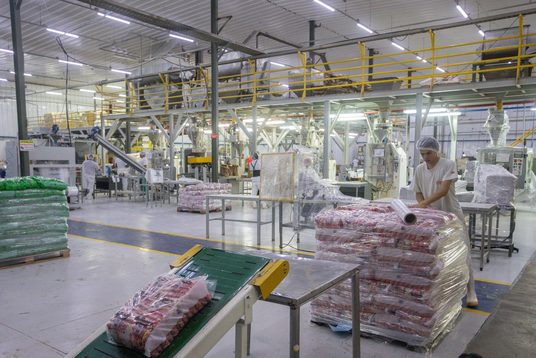 Trabalhador embrulha palete de produtos embalados com filme plástico em galpão fabril amplo.