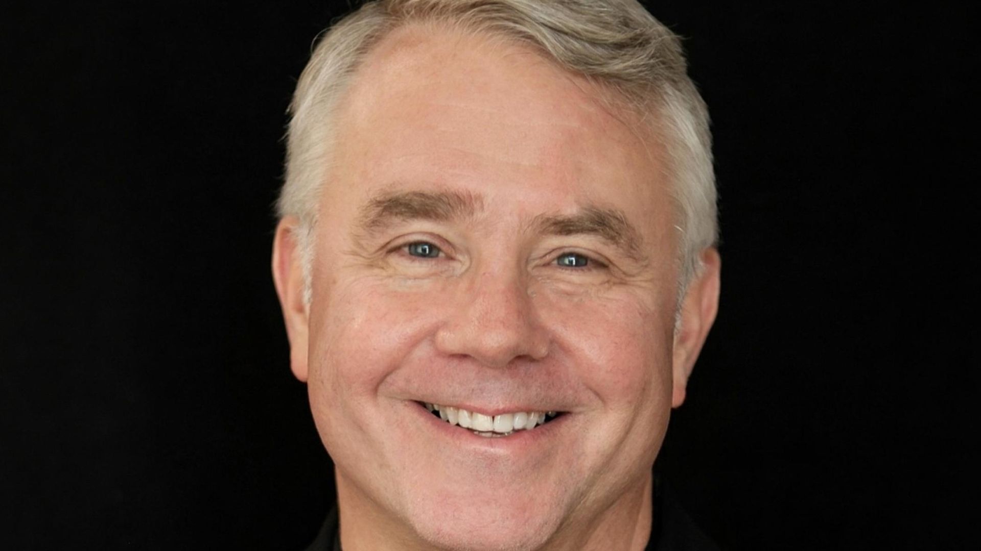 Um close do rosto de John Warner, um homem mais velho de cabelos grisalhos e olhos azuis, sorrindo calorosamente contra um fundo preto sólido. Ele tem rugas sutis de riso ao redor dos olhos e boca.
