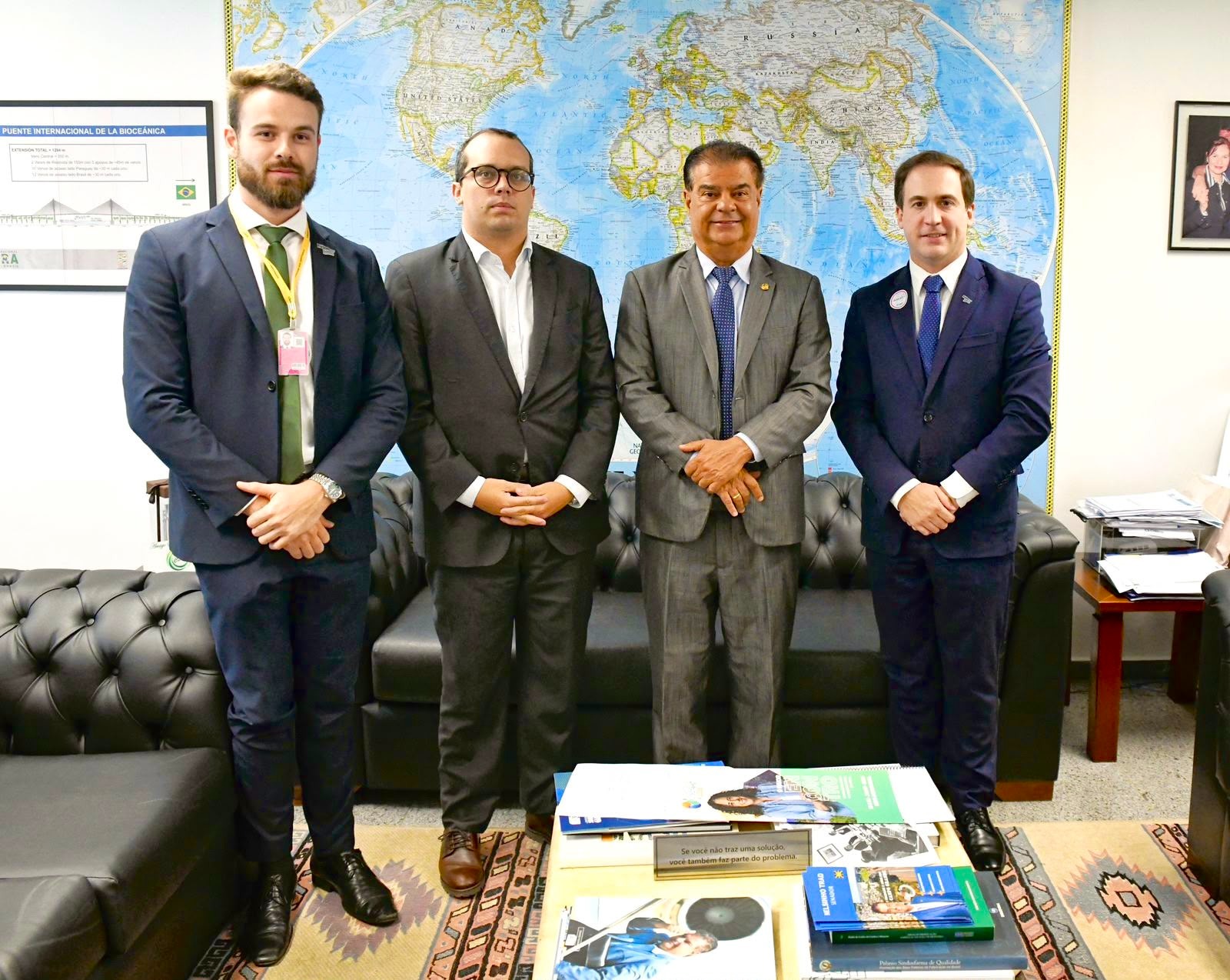 Quatro homens de terno alinhados em pé durante reunião em um escritório. Ao fundo, um mapa-múndi na parede.