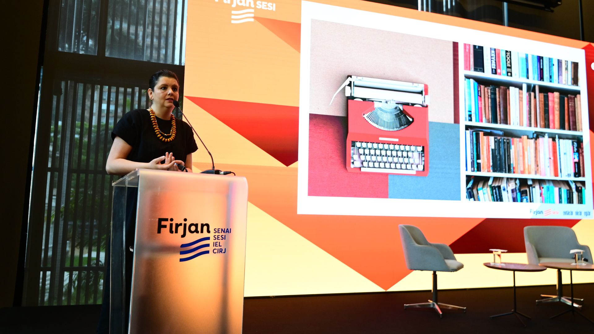 Mulher discursa em púlpito com a marca Firjan durante evento. Ao fundo, telão exibe apresentação com imagens de uma máquina de escrever e uma estante de livros.