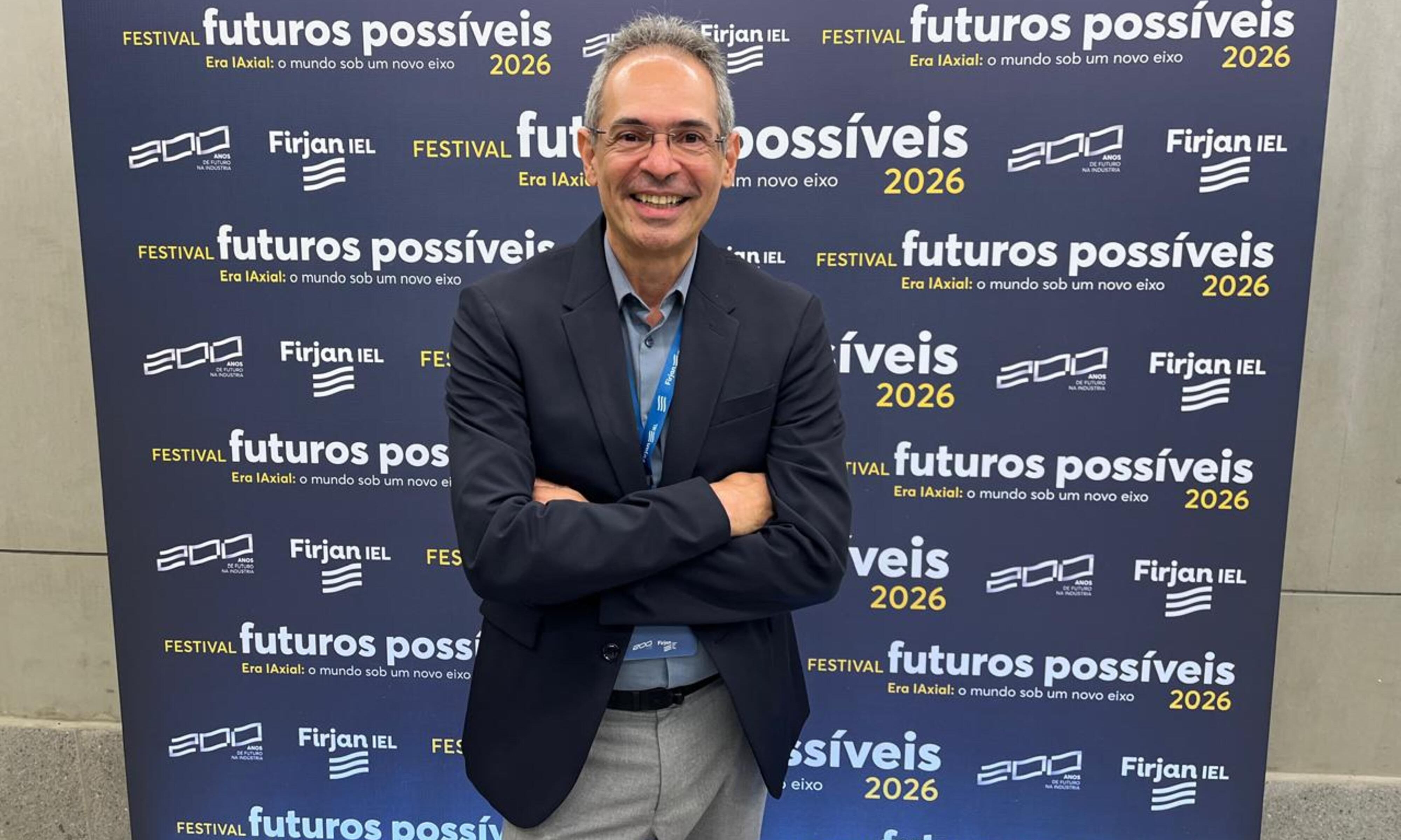 Luiz Mandarino sorri de fato e camisa azul diante do painel do Festival Futuros Possíveis 2026 da Firjan IEL.